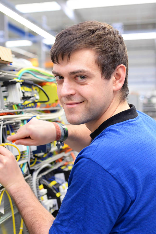 Junger Industrieelektriker Lehrling mit blauen Shirt baut gerade an einer Baugruppe und lächelt in die Kamera